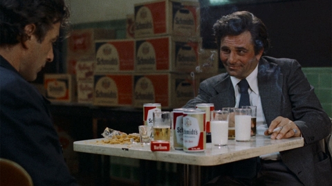 Two men sit at a table with cans of Schmidt's beer, glasses of milk and beer, a cigarette, and snacks in a dimly lit room.