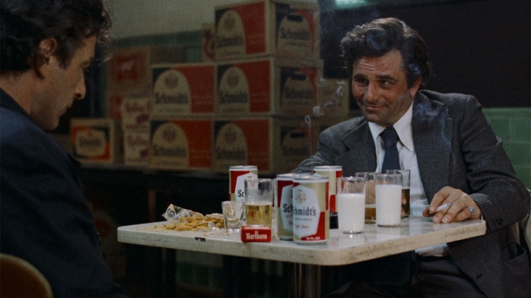 Two men sit at a table with cans of Schmidt's beer, glasses of milk and beer, a cigarette, and snacks in a dimly lit room.