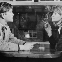 Black and white photo of two people sitting at a table across from each other, with one person smoking a cigarette indoors.