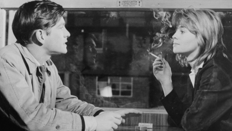 Black and white photo of two people sitting at a table across from each other, with one person smoking a cigarette indoors.