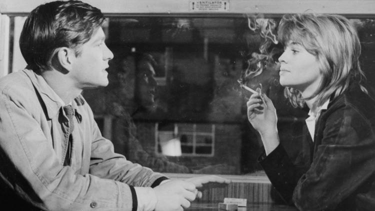 Black and white photo of two people sitting at a table across from each other, with one person smoking a cigarette indoors.