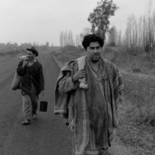 A still from The Jackal of Nahueltoro of two men walking on a rural road, carrying belongings, with leafless trees and overcast skies in the background.