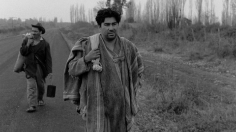 A still from The Jackal of Nahueltoro of two men walking on a rural road, carrying belongings, with leafless trees and overcast skies in the background.