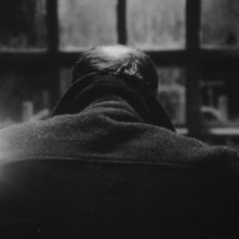 Black and white photo of a person with thinning hair sitting at a desk facing a window with a bright lamp on the left.