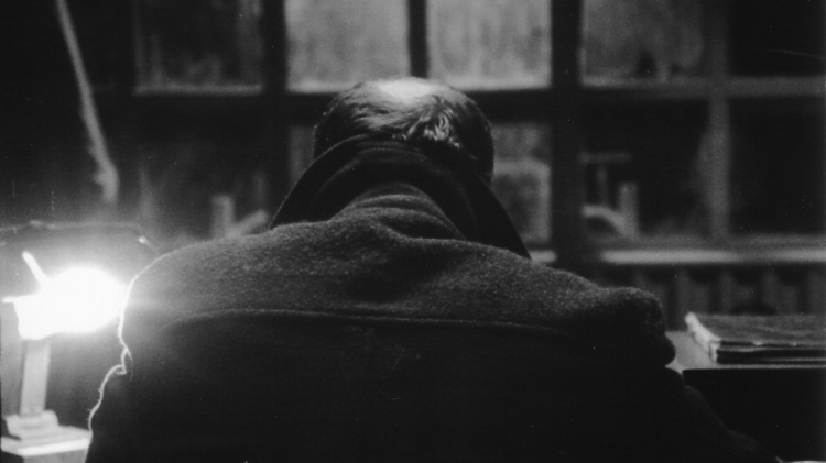 Black and white photo of a person with thinning hair sitting at a desk facing a window with a bright lamp on the left.