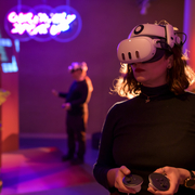 Person wearing a VR headset holds controllers in a dim room with neon lights. Another person in the background also uses VR, creating a futuristic ambience.