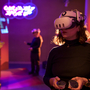 Person wearing a VR headset holds controllers in a dim room with neon lights. Another person in the background also uses VR, creating a futuristic ambience.