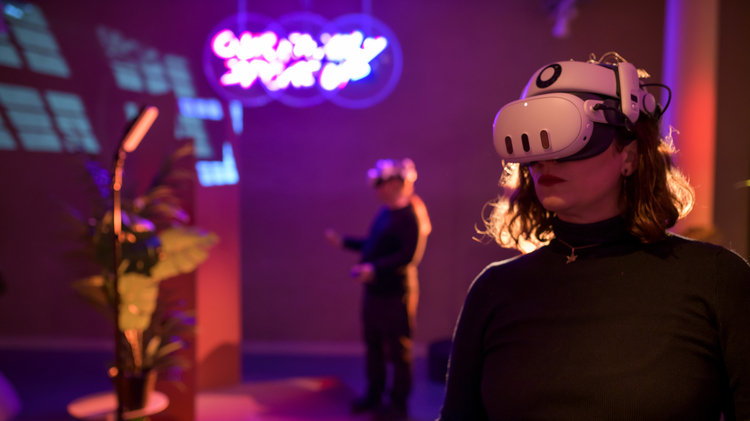 Person wearing a VR headset holds controllers in a dim room with neon lights. Another person in the background also uses VR, creating a futuristic ambience.