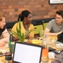 A group of five people sits around a table engaged in discussion. Laptops, notebooks, and drinks are on the table, creating a collaborative atmosphere.