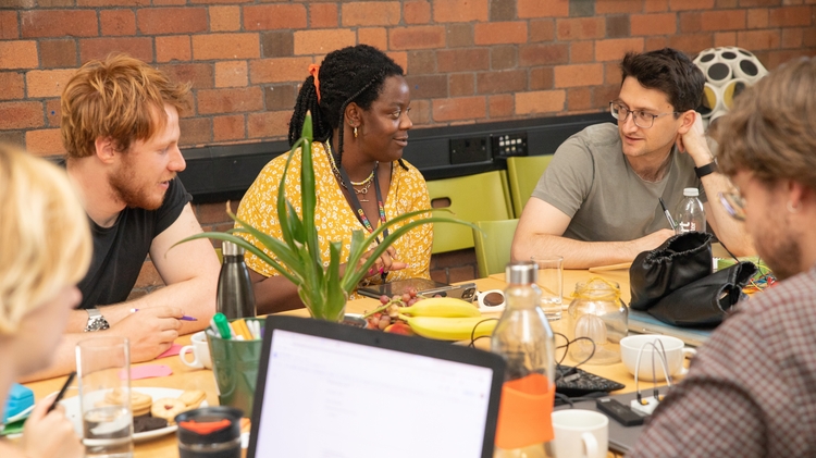 A group of five people sits around a table engaged in discussion. Laptops, notebooks, and drinks are on the table, creating a collaborative atmosphere.