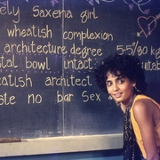 Person in yellow sitting beside a blackboard with handwritten text describing a "Homely Saxena girl" and personal details.