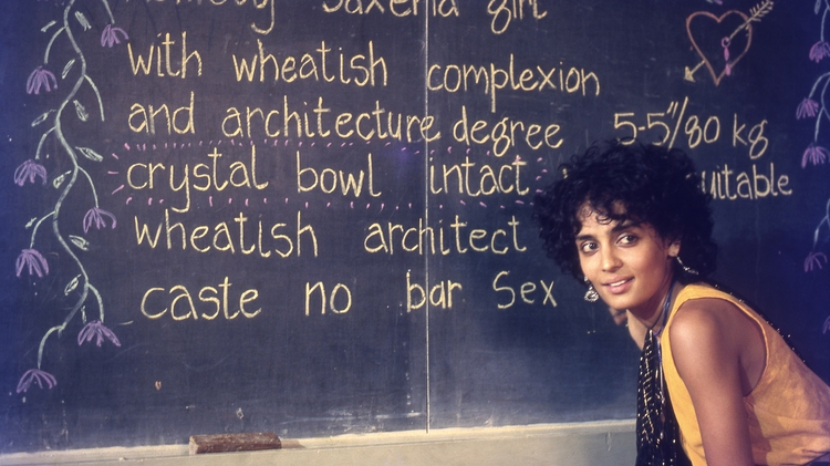 Person in yellow sitting beside a blackboard with handwritten text describing a "Homely Saxena girl" and personal details.