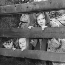 A black and white photo of three children looking through a fence.