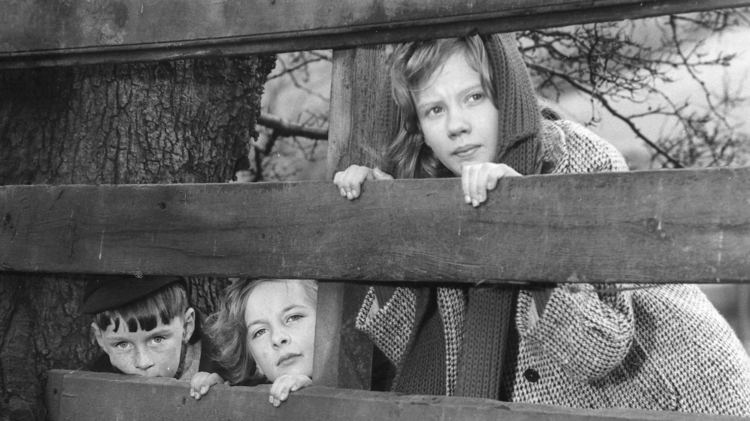 A black and white photo of three children looking through a fence.