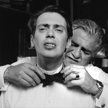 Black and white photo of an older man helping a younger man adjust a bow tie in a kitchen setting.
