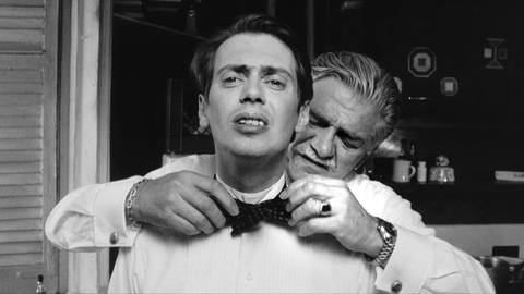 Black and white photo of an older man helping a younger man adjust a bow tie in a kitchen setting.