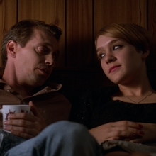 Two people sitting closely on a couch against wooden paneling, one holding a mug, in a dimly lit room.