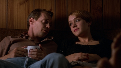 Two people sitting closely on a couch against wooden paneling, one holding a mug, in a dimly lit room.