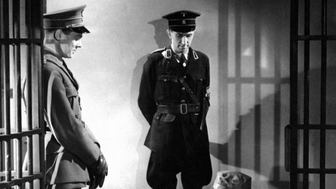 Black and white photo in a jail setting of two uniformed guards looking down at a prisoner on their knees.