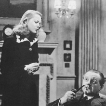 A woman looks down at a man who is seated smoking a pipe in a grand living room setting.