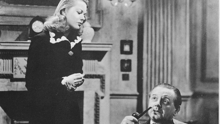 A woman looks down at a man who is seated smoking a pipe in a grand living room setting.