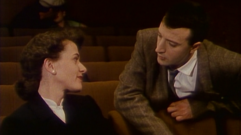A photo of a man leaning over a cinema seat to talk to a woman.