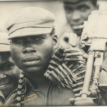 A still of a man holding a gun looking at the camera with ammunition wrapped around his neck.