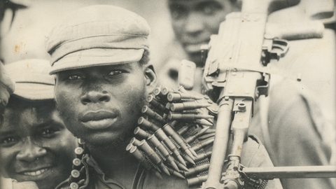 A still of a man holding a gun looking at the camera with ammunition wrapped around his neck.