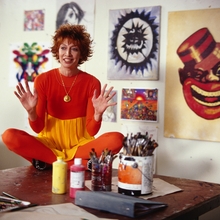 A woman in an all-orange outfit sits cross-legged on a table with paintbrushes and paint, surrounded by artwork on the walls.