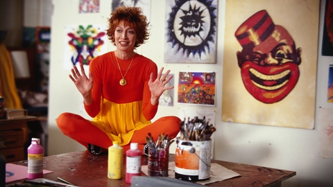 A woman in an all-orange outfit sits cross-legged on a table with paintbrushes and paint, surrounded by artwork on the walls.