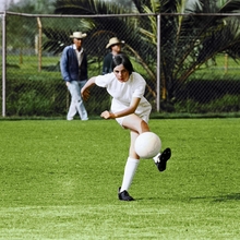 Young soccer player kicking ball on green field with spectators and palm trees in background.