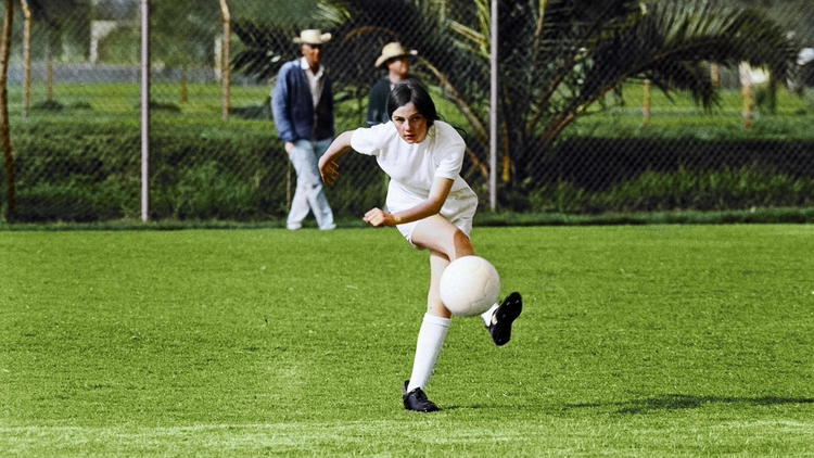 Young soccer player kicking ball on green field with spectators and palm trees in background.