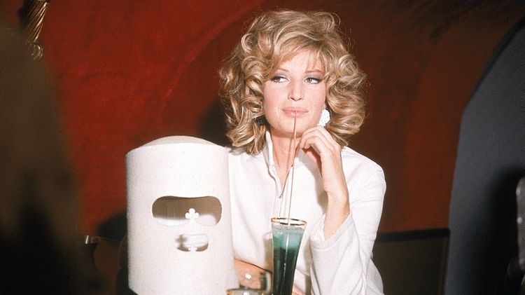 Woman with curly blonde hair in a white outfit seated at a bar table with multiple drinks.