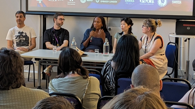A panel of people sitting in front of an audience with a screen behind them.