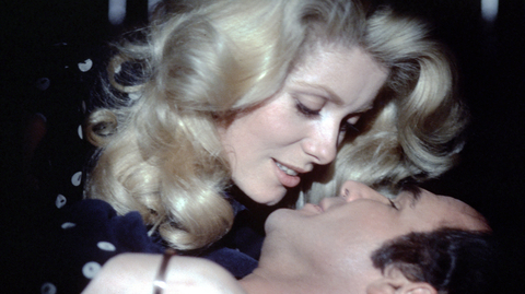 A close-up of a man and a woman embracing and leaning in for a kiss in a dimly lit setting.