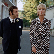 Two people in formal attire walking on a quiet street lined with greenery and a hotel restaurant sign in the background.