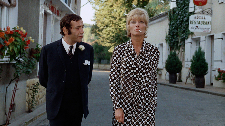 Two people in formal attire walking on a quiet street lined with greenery and a hotel restaurant sign in the background.