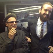 Two men posing indoors; one wears a hat and coat, the other wears a suit with a floral tie, standing in front of a poster with a white car.