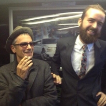 Two men posing indoors; one wears a hat and coat, the other wears a suit with a floral tie, standing in front of a poster with a white car.
