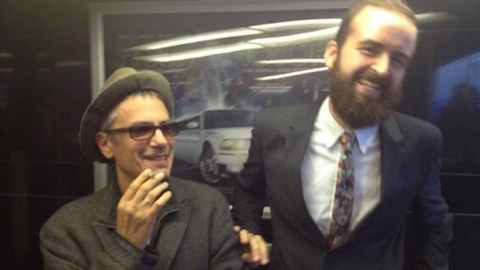 Two men posing indoors; one wears a hat and coat, the other wears a suit with a floral tie, standing in front of a poster with a white car.