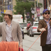 Two men dressed in casual blazers handling a reel of orange cable on a city street with cars and a streetcar in the background.