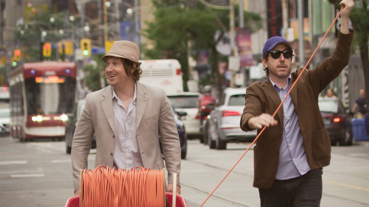Two men dressed in casual blazers handling a reel of orange cable on a city street with cars and a streetcar in the background.