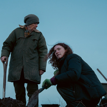 Two people in winter clothing working with shovels and a wheelbarrow in a field under a clear blue sky at dusk.
