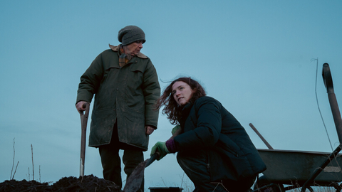 Two people in winter clothing working with shovels and a wheelbarrow in a field under a clear blue sky at dusk.