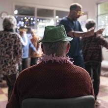 Elderly person wearing a green party hat watches a lively holiday gathering with dancing and festive decorations in a cozy room.