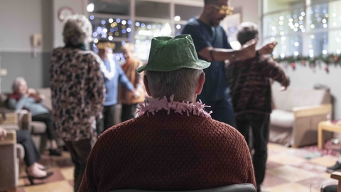 Elderly person wearing a green party hat watches a lively holiday gathering with dancing and festive decorations in a cozy room.