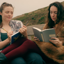 Two people sit on a bench outdoors; one writes in a notebook while the other reads a book, surrounded by grassy hills.