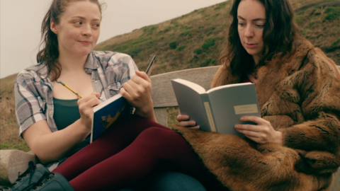 Two people sit on a bench outdoors; one writes in a notebook while the other reads a book, surrounded by grassy hills.