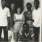 Black-and-white photo of six people, five standing and one sitting, posing casually in front of a plain wall and door.