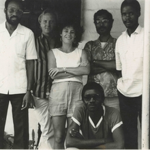 Black-and-white photo of six people, five standing and one sitting, posing casually in front of a plain wall and door.